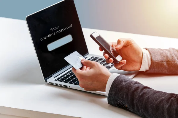 A young man in a suit pays for the purchase in an online store, holds a phone and credit card in the background of a laptop. Hands close-up on a blue background