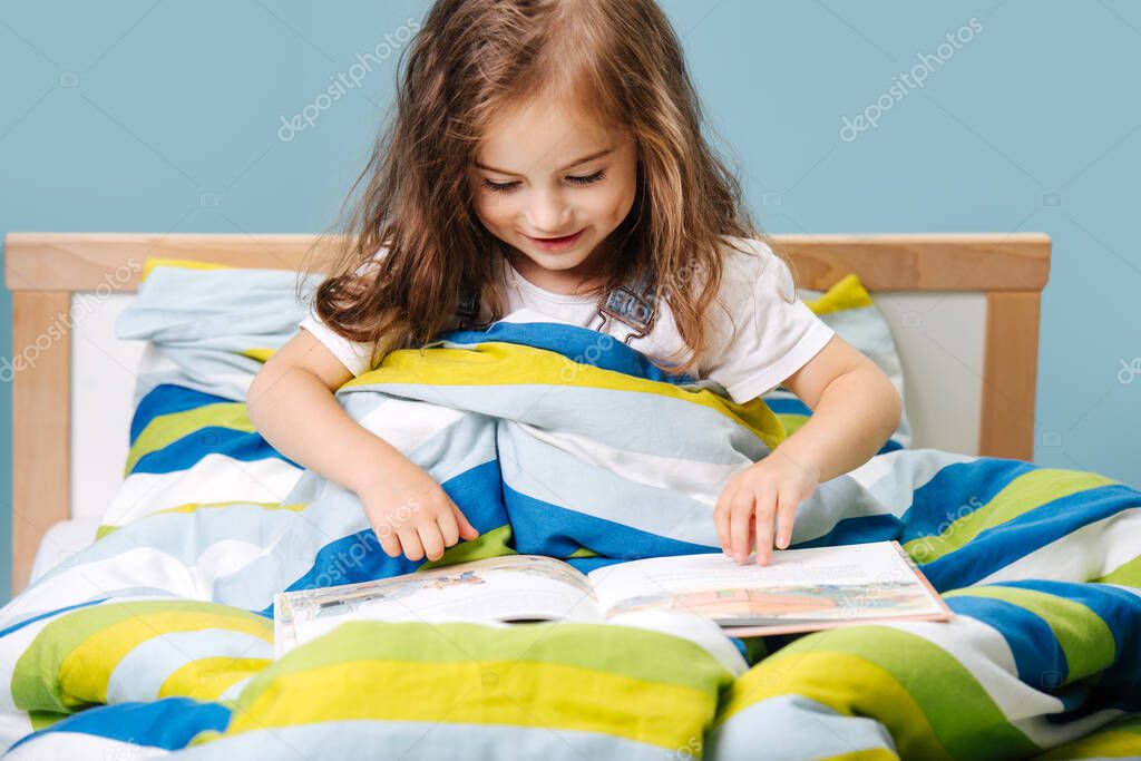 Girl Toddler with curly hair prepares for bed and flips a children's book sitting in bed on a blue background