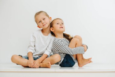 Masada bacakları havada oturan yalınayak komik küçük kardeşler. Her ikisi de kot şort ve uzun kollu giyiyor. Çocuk rahat bir şekilde oturuyor ve kız ona yaslanıyor, dili dışarı çıkıyor..