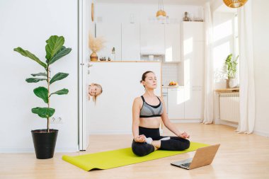Sarışın bir çocuk odaya gizlice bakıyor, annesi bilgisayarın önünde yoga kursuna gidiyor, lotus pozisyonunda oturuyor ve beyaz bir mutfağın arka planında meditasyon yapıyor.