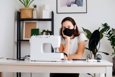 Evdeki bir masada elinde dikiş makinesiyle oturan bir kadının portresi, siyah kumaştan koruyucu bir maske dikmiş ve onu denemiş.