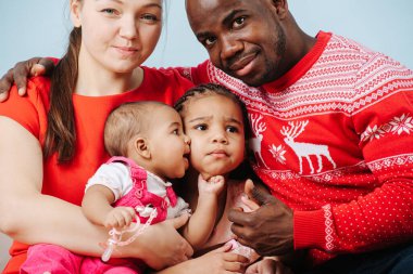 Kırmızı elbiseli mutlu melez aile bir stüdyoda aile fotoğrafı için poz veriyor. Siyah baba, beyaz anne ve iki küçük kızı mavi arka planda..