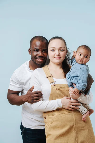 Happy mixed race family posing for a family photo in a studio. Black father, white moher and their infant daughter over blue background. All looking at camera.