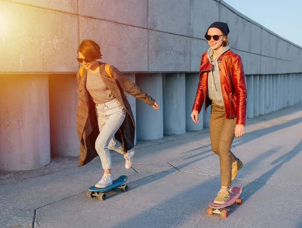 サングラスをかけた二人の若い幸せな女性がスケートボードに乗っている 街の夕日 灰色の床と建物 速度を上げる — ストック写真