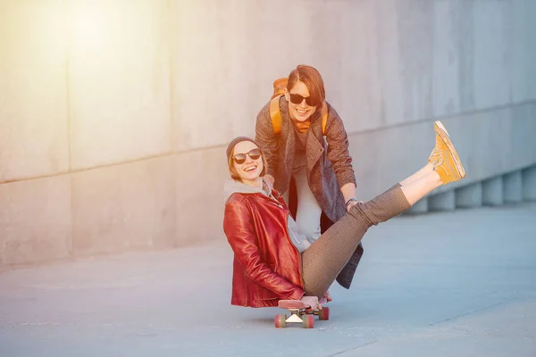 サングラスで短い髪を持つ2人の若い面白い女性の肖像画 そのうちの1人は 空中に彼女の足でスケートボードに座っています 2つ目は 速度を上げるためのプッシュを与えている — ストック写真