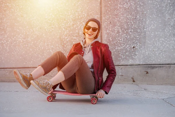赤いジャケット タイトなブリーチとサングラスで短い髪を持つ若い面白い女性の肖像画 彼女はスケートボードの上に座ってる — ストック写真