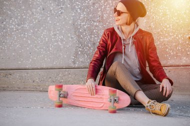 Kırmızı ceketli ve güneş gözlüklü kısa saçlı mutlu bir kadının portresi. Bir sokak zemininde oturuyor. Bir elinde pembe kaykayını tutuyor. Dişleriyle gülümsüyor.