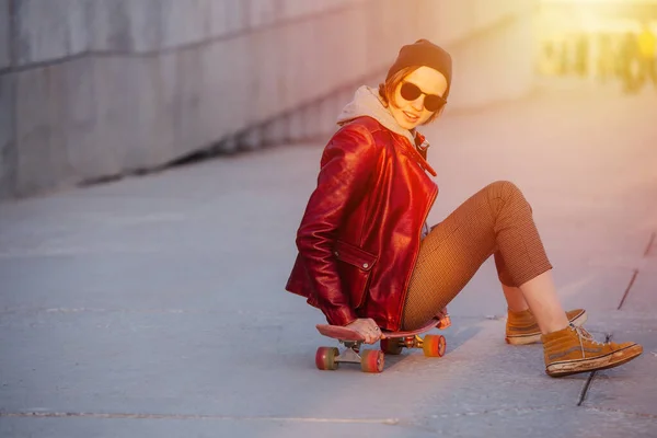 赤いジャケット タイトなブリーチとサングラスで短い髪を持つ若い面白い女性の肖像画 彼女はスケートボードに座っているこの面白い位置に乗って — ストック写真