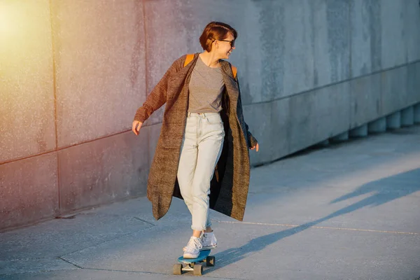 日没の乗馬スケートでライトグレーのファッショナブルなコートで短い髪を持つ若い幸せな美しい女性の肖像画 彼女は動きながら肩の後ろを見てる — ストック写真
