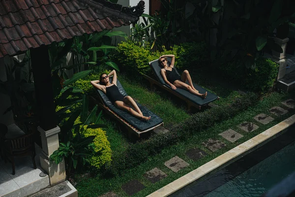 黒の水着を着た2人の若い女性と トロピカルリゾートハウスの中庭にあるデッキチェアでリラックスしたサングラス 高い角度だ 太陽を楽しみ リラックス — ストック写真