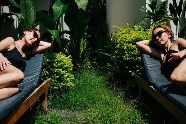 两名身穿黑色泳衣 戴着太阳镜的快乐的年轻女子在热带度假别墅的庭院里 坐在躺椅上休息 放松一个在另一个前面 低角度 复制空间 — 图库照片