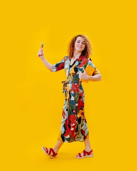 黄色の上にノートやクレヨンで芸術的に歩く幸せな女性デザイナー 笑顔でカメラを見て — ストック写真