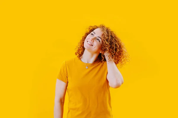 Half-length image of a cheerful woman with curly red hair in an orange shirt over yellow background. Looking up, daydreaming about something positive.