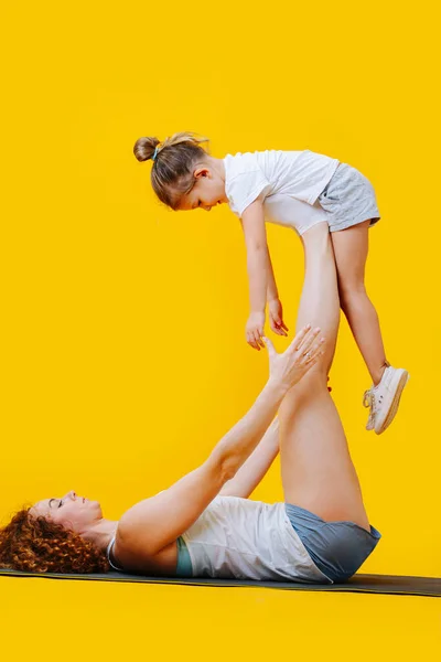 黄色の背景にヨガマットの上の母親と娘の体操フォーム まっすぐ足の上で空気中の女の子を保持女性 — ストック写真
