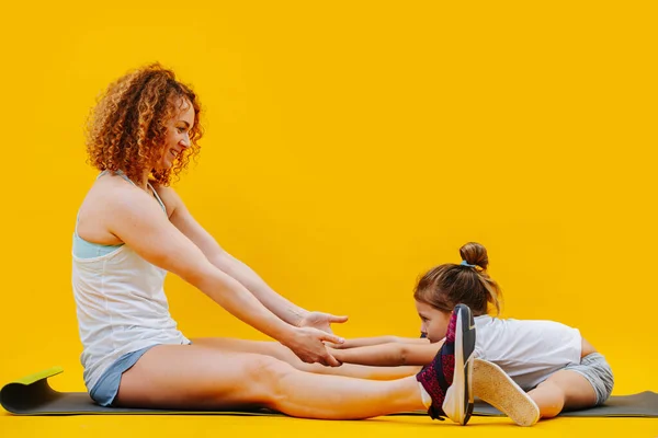 妈妈和女儿在做体育锻炼 在黄色的背景上同步 压着脚 拉着手往下弯曲 伸展大腿内侧肌腱 — 图库照片