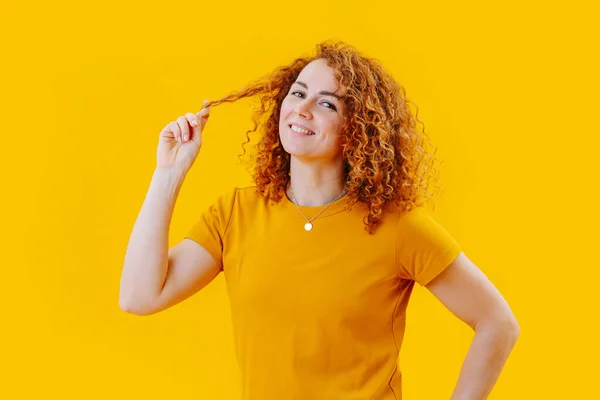 Счастливая молодая женщина играет с кудрявыми рыжими волосами над желтыми, глядя в камеру — стоковое фото