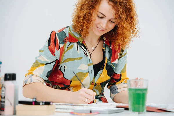 Sorrindo artista mulher pintura atrás de mesa de trabalho em seu bloco de notas com uma escova — Fotografia de Stock