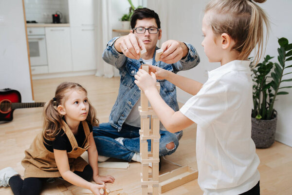 Dad playing with kids on the kitchen floor, helping his little son and daughter build tower from wooden blocks. Spending time with kids on isolation.