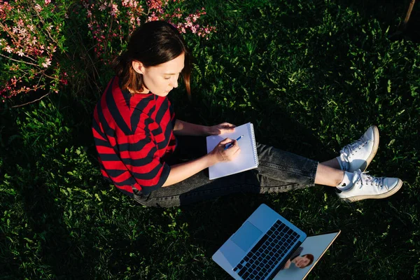 Top view of girl sitting on the grass in a flowering garden studying online with laptop
