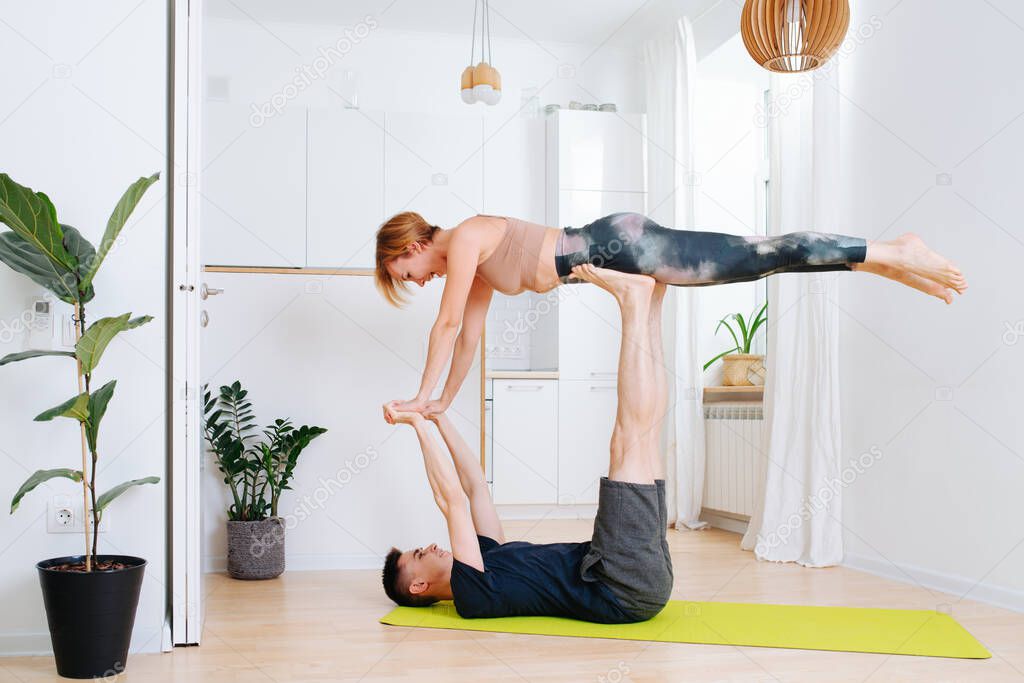 Adult siblings doing gymnastics together during isolation at home. Guy lying on his back on a mat, holding girl in the air with straight legs and arms