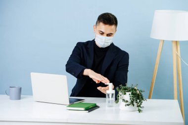 Covid-19 salgını sırasında bir ofiste çalışan maskeli bir adam. Çalışma masasındaki hiçbir şeye dokunmadan önce ellerini dezenfekte ediyor..