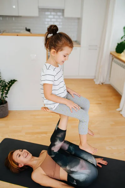 Young mom doing gymnstics with her little daughter on a yoga mat at home. Mom lays on her back, girl sitting on her raised feet like on the chair.