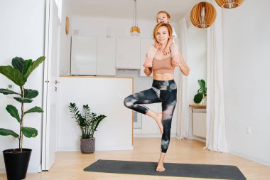 Annesi evde minderde yoga yaparken küçük oğlu omuzlarında oturuyordu. Sıkı spor kıyafetler giyiyor.