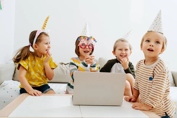 Four little kids having online birthday party on a quarantine at home. They are celebrating in front of laptop, wearing party cone hats.