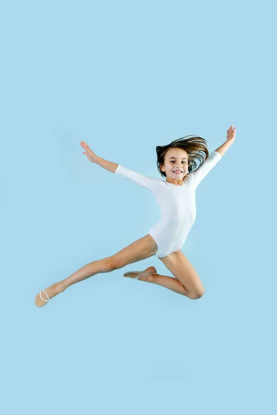 穿着白色紧身衣的黑发小女孩以体形做体操 跳过蓝色的背景 高兴地看着摄像机 — 图库照片