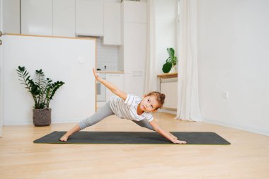 Evdeki minderde yoga yapan küçük kız. Bacakları ayrık bir şekilde öne eğiliyor, bir tarafı düz bir şekilde yere değiyor, diğer tarafı koluyla kaldırıyor..