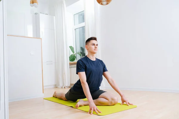 Tall man doing yoga on a mat in a kitchen at home during isolation. Lifting upper body with hands, with legs on a mat. Leg behind is straight, leg in front is folded.