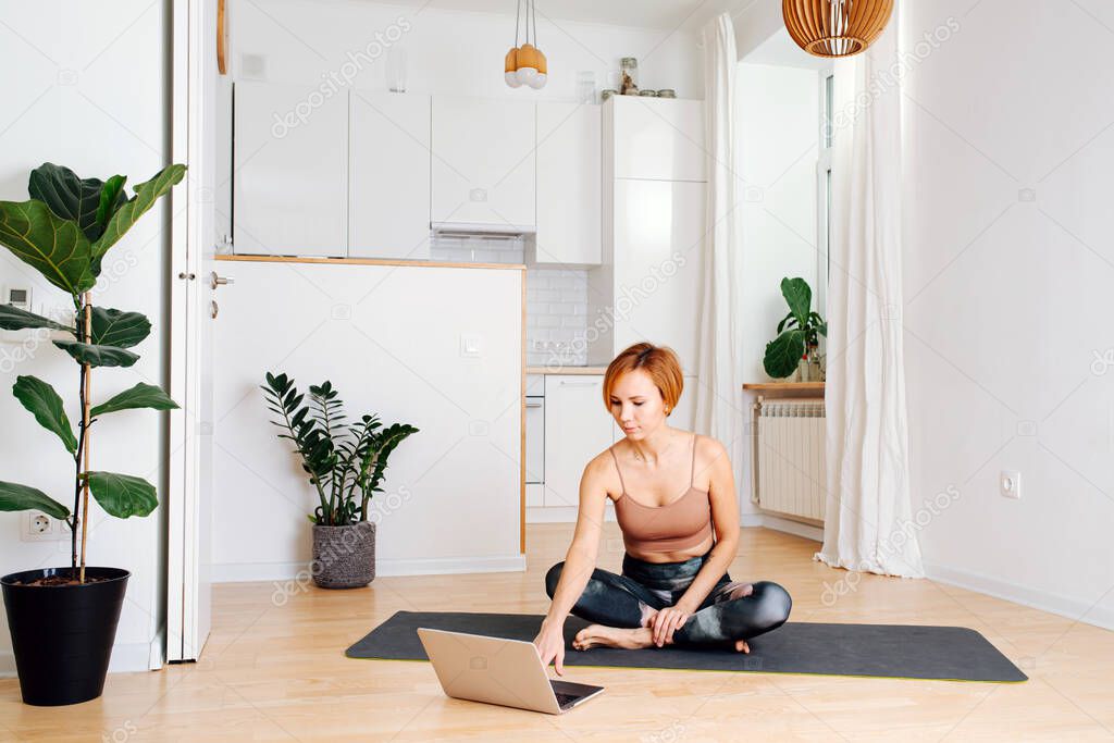 Woman doing yoga on a yoga mat in front of a laptop at home. She wears tight sportive clothes. Choosing appropriate track.