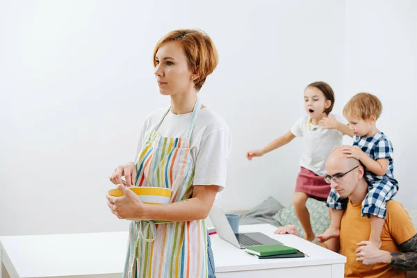 Tired family during covid-19 isolation. Mom cooking in apron, kids watch dad work on his laptop.