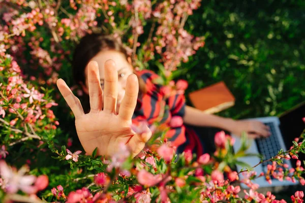 Young woman sitting with laptop in the bush blooming with pink flowers. She\'s blocking camera with her hand. top view.