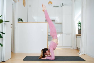 Yogi kadını yoga yapıyor, pembe spor giysisi giyiyor, salamba sirsasana egzersizi yapıyor, uzun amuda kalkıyor, evde çalışıyor.