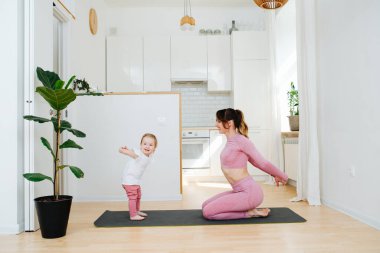 Spor giyim sektöründe genç bir anne evde oturup bebeğiyle egzersiz yapıyor, ellerini arkasından tutuyor ve esneme hareketleri yapıyor.