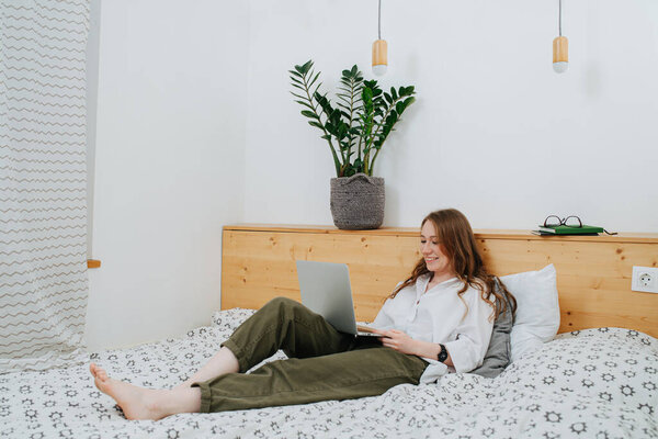 Счастливая молодая женщина сидит в двуспальной кровати с ноутбуком, улыбаясь
