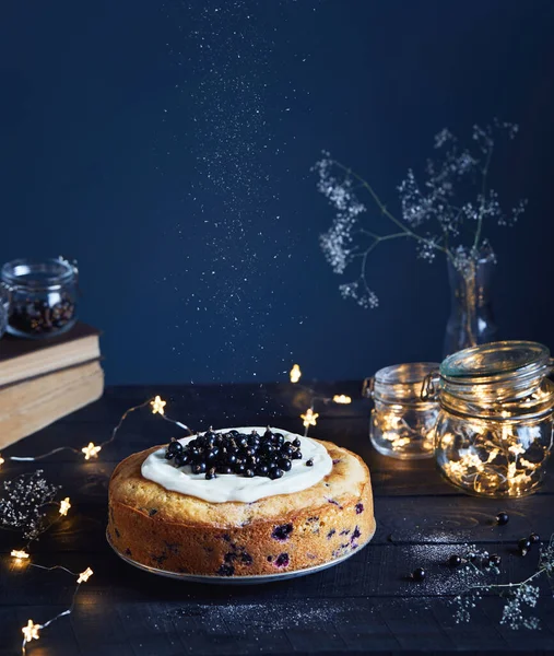 Homemade cake with black currant and mascarpone on the wooden table near old books and glass jars with light stars on dark background in summer time