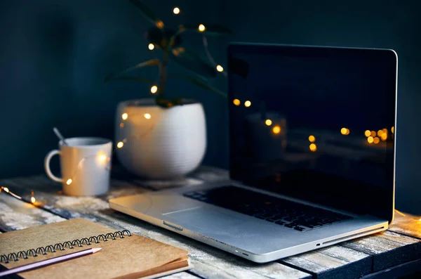 Cozy workspace with laptop, note with pencil and cup of coffee, plants and yellow glowing garland at stone grey wall background. Working from home concept.