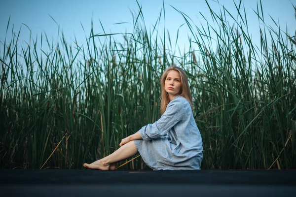 Romantic girl in blue dress seating on pier. Vegetative background — Stock Photo, Image