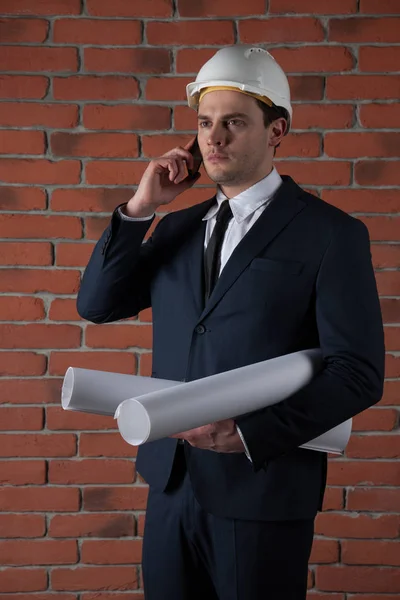 Retrato de um homem de negócios com smartphone em capacete branco no fundo da parede de tijolo vermelho — Fotografia de Stock