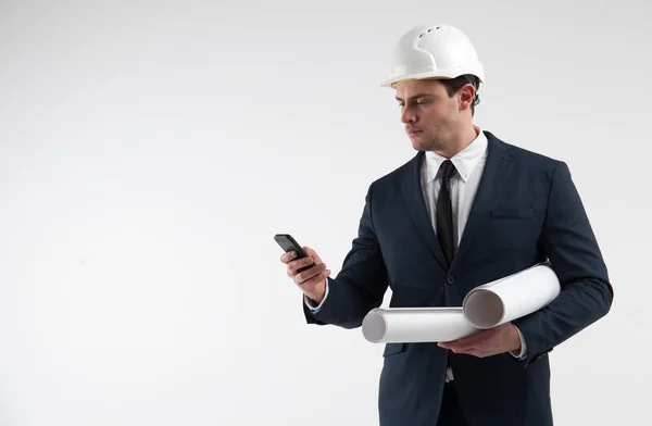 Retrato de um arquiteto no capacete branco olha para o smartphone no fundo cinza claro — Fotografia de Stock