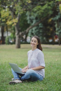 Laptoplu genç bir kadın yeşil çimlerde oturuyor.