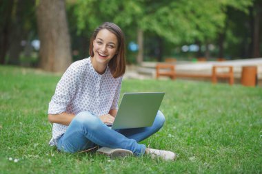 Laptoplu genç bir kadın yeşil çimlerde oturuyor.