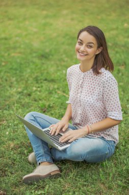 Laptoplu genç bir kadın yeşil çimlerde oturuyor.