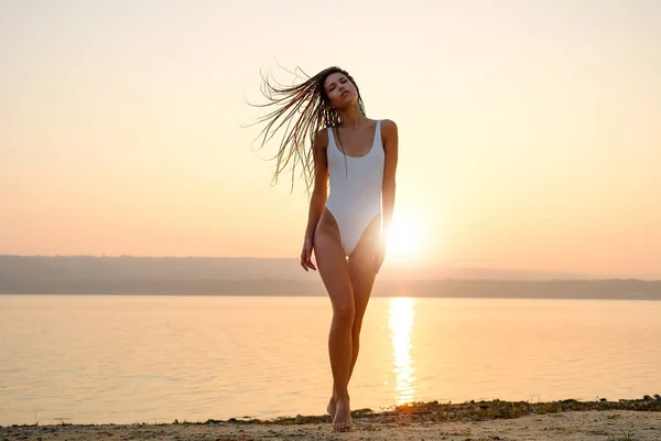 Mujer joven en traje de baño de cuerdas se para en la playa al amanecer — Foto de Stock