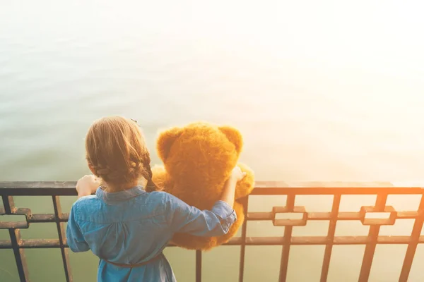 Girl embracing a cute teddy bear looking to water — Stock Photo, Image