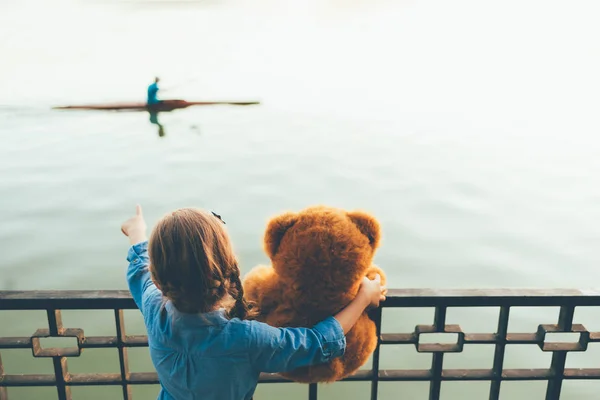Visão traseira da menina abraçando um urso de pelúcia bonito mostrando a uma canoa — Fotografia de Stock