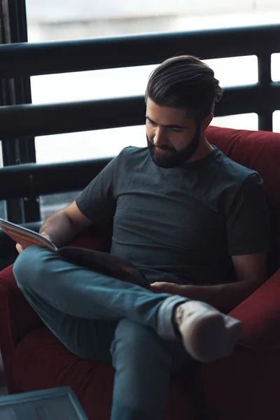 Retrato hombre barbudo guapo con ropa casual, sentado en la silla roja estudio loft moderno — Foto de Stock