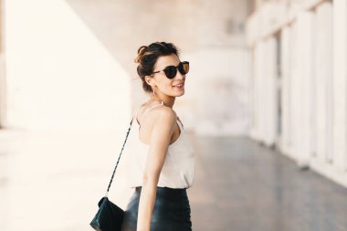 Güneş gözlüklü güzel, gülümseyen genç bir kadın yürürken omzunun üstünden bakıyor.